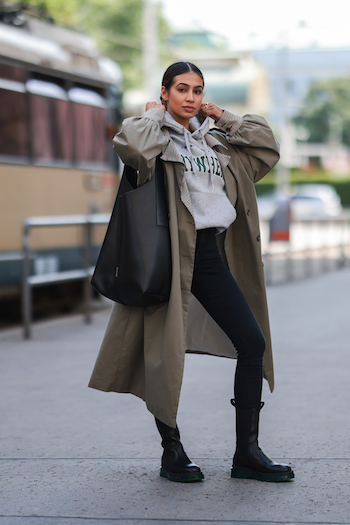 Sonia Dhillon wearing a khaki trenchcoat, a black denim jeans, a grey hoodie with green print on it, a copenhagen Bag 1 in vitello black and the CPH1000 in vitello black/green. She is standing on the streets of Vienna. Behind her you can see a train.