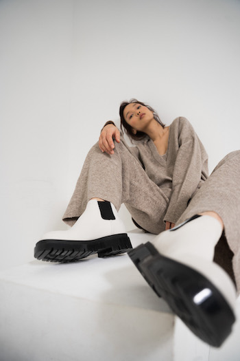 Picture from the Lookbook shooting of the Autumn Winter Collection 2021 from Copenhagen Studios. Model is sitting on a white block and stretch out one leg. She is wearing a beige cashmere set. The outfit is combined with the CPH1001 Boot in Vitello Eggshell.