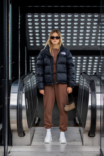 @alexawinter is standing in front of the escalator. She wears a dark brown loungewear suit in combination with a black puffer jacket from The North Face, the CPH40 in vitello white and cool black sunglasses.