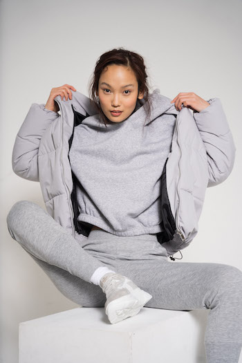 Campaign Picture from the Lookbook Shooting Autumn Winter 2021 from the brand Copenhagen Studios. Model is sitting on a white block and is wearing a grey monochrome outfit with CPH51 Material Mix Sneakers. The Outfit consists of a grey leggings, a grey hoodie and a grey puffer jacket.