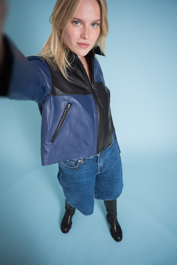Campaign picture from the tone in tone shooting from Copenhagen Studios. Model is standing and pretending to do a selfie with the camera. She is wearing knee length pants a black and blue leather jacket and the CPH551 Vitello black knee boots.