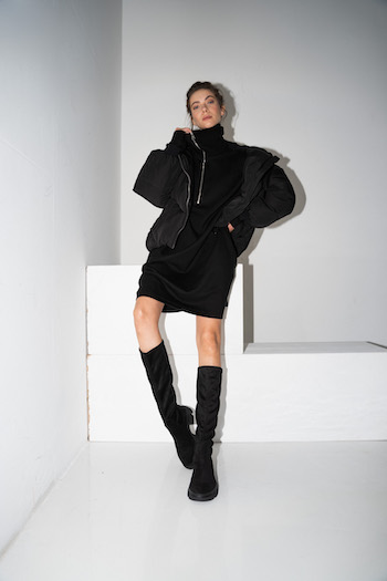 Picture from our Lookbook Shooting Autumn Winter 2021. The model leans against a white lock. She is wearing a black oversized sweater dress with a black puffer jacket and the Copenhagen cph556 costa black kneeboots.