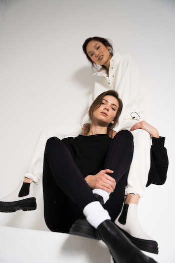 Lookbook Picture from the Autumn Winter Collection 2021 of Copenhagen Studios. Two models are sitting. One is sitting higher on a block. Picture is made from a low perspective. One model is wearing a white jogger set with the CPH1001 Eggshell and the other model is wearing a black sweater and black leggings with the CPH1001 in Vitello black with white tennis socks.