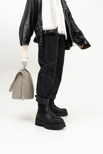 Cph686 Vitello Black. Model is holding a bag in the color stone in her hand. She wears black boots with a high plateau sole. The model wears a black leather jacket and a black mom jeans.