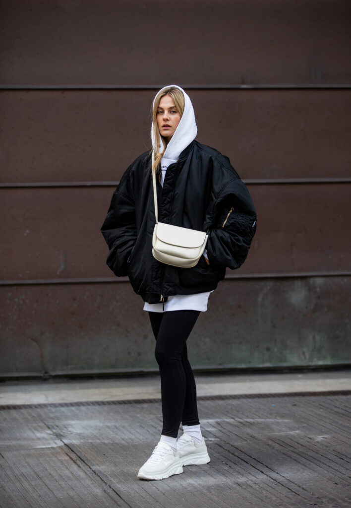 During the Fashion Week in Copenhagen 2022 the sister Anna winter and Alessa Winter did a street style shooting with Copenhagen Studios Sneakers. On this picture you can see Alessa Winter wearing CPH64 material mix white with a leggings, an oversized hoodie and a bomber jacket. She stand infant of a brown wall.
