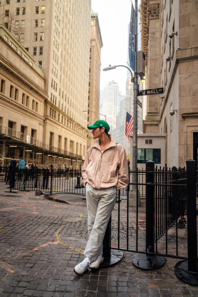 Jonas Kautenburger standing on the streets of New York City. He wears his  Copenhagen Studios sneaker CPH350M calf white/black in combination with a light grey chino trousers, a pastel pink zip top and a bright green New Era cap.
