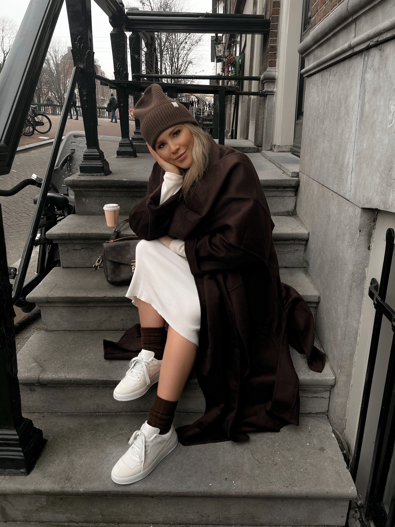 @verena_ahmann is wearing a outfit in natural tones. She combined her CPH461 leather mix cream with a beige silk dress, a dark brown oversized trenchcoat, dark brown socks and a beanie. She is sitting on some stairs in Amsterdam.