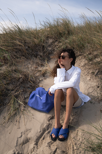 Shooting from the THOC. The influencer Joellejuana is sitting at the beach with her new CPH Sandals in blue. She is wearing a white oversized blouse.