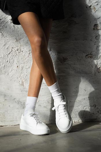 The classic white sneaker CPH407 vitello white. The model is laying against a wall wearing white sneakers with white socks and a black sporty shorts.