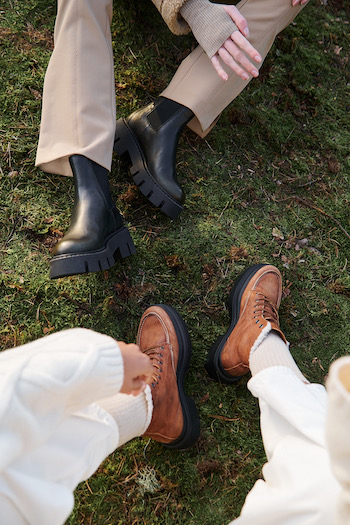 Our new styles in Studio Lilo’s installation. Two girls sitting on the ground on a green moss installation. One model is wearing a black boot and the other one a brown hiking boot.