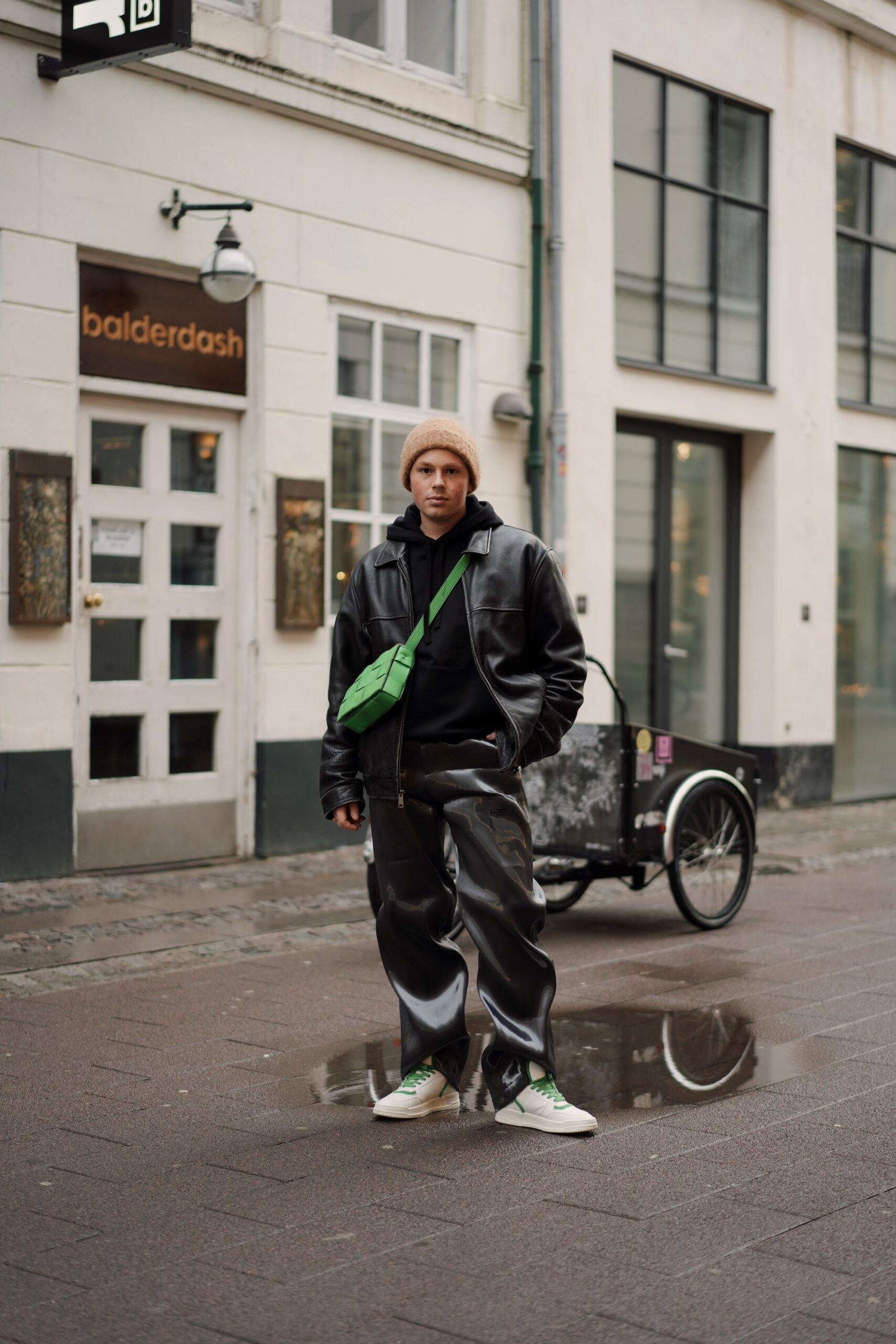 The young male Influencer Erik Scholz is standing on the streets of Copenhagen. He is wearing an combination of leather trousers and a leather Bomberjacket both in black. He combined his outfit with our latest sneakers, the CPH196 vitello white green and a green bag.