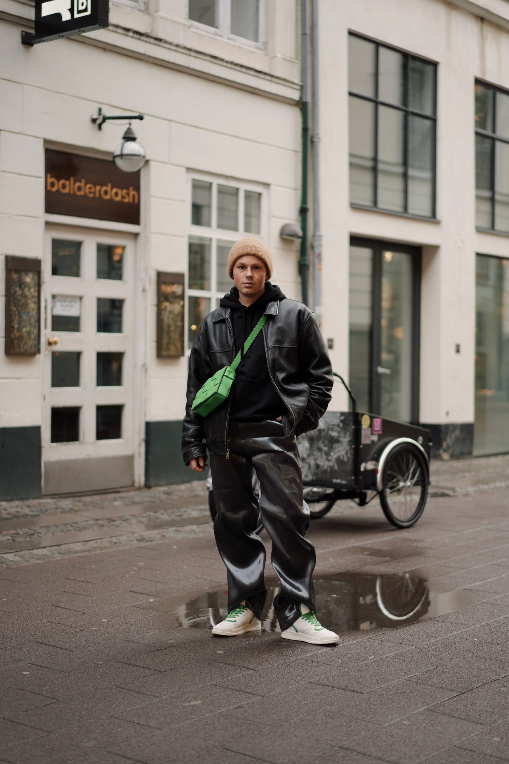 The young male Influencer Erik Scholz is standing on the streets of Copenhagen. He is wearing an combination of leather trousers and a leather Bomberjacket both in black. He combined his outfit with our latest sneakers, the CPH196 vitello white green and a green bag. 