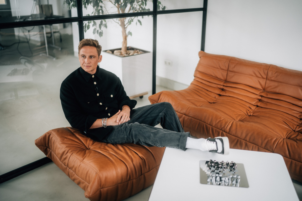 Matthias Schweighöfer, a well-known German actor is setting on a brown leather sofa in his office space in Berlin-Mitte- He is wearing the new Copenhagen Studios Sneaker CPH1M in vitello black. His feet are laying on the side table of the are.  