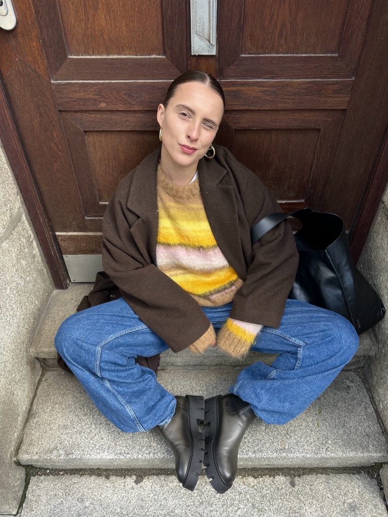 The young creator Lara Bussmann from duesseldorf is sitting on stairs in front of a wooden door wearing our Vintage Boot CPH137. She is also wearing a yellow striped wool hoodie, blue denims and a brown coat.