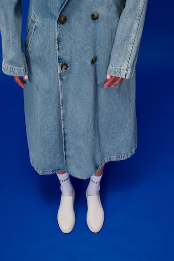 The white sneaker CPH204 in white worn by a model wearing a denim skirt. She is standing on a blue backdrop with her hands down.