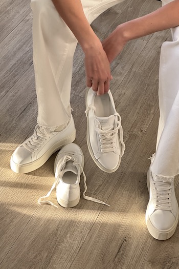 Vera Van Erb holding our CPH209 leather milk in one hand & the CPH4 soft vitello white in the other. She is sitting, wearing beige trousers.