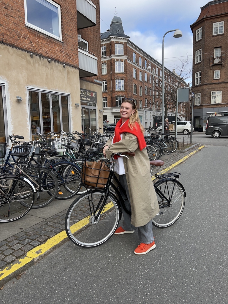 Maria Jonsson posing with her black bike, wearing our CPH213 suede royal red sneaker combined with a beige trenchcoat, grey jeans, a white shirt & a matching red pullover.