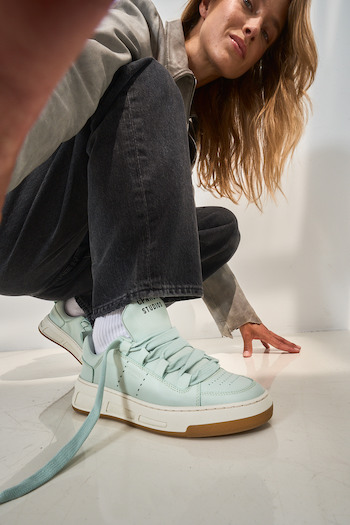 A female, blonde model is wearing our CPH213 soft vitello sage, taking a selfie while putting the shoe is focus. 