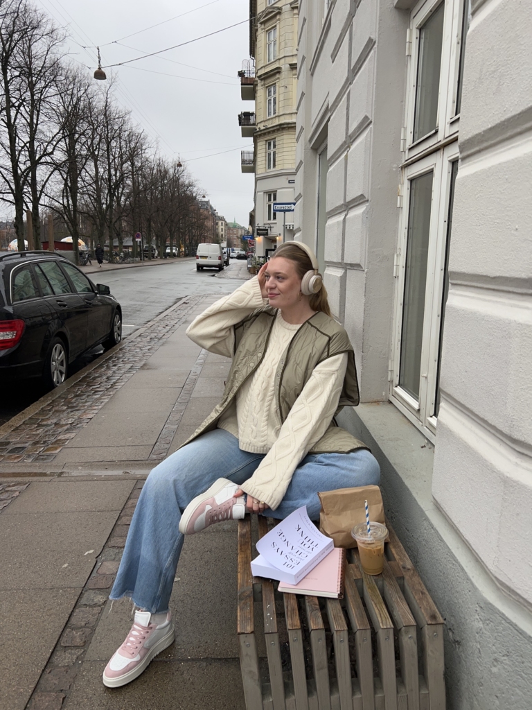 Maria Jonsson sitting on a bench wearing her CPH77 leather mix white/rose shoes together with blue jeans, a beige pullover, a vest & headphones.